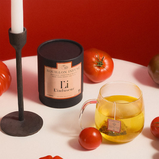 Candle with ' L'infuseur' packaging, glass mug with yellow liquid, and tomatoes on a white surface against a red backgroundm Organic Tomato–Celery Broth by L'Infuseur – Vegetable Blend