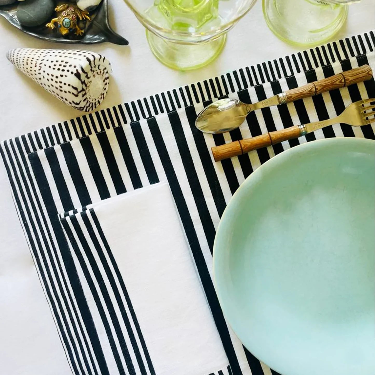 Table setting with black and white striped placemats, a light blue bowl, and silverware on a white surface. Milleraies Striped Linen Placemat – Aurelie Bidermann – Maison Holder