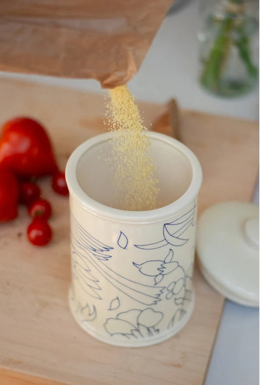 Ceramic container with illustrations being filled with a yellow powder on a wooden surface. Rouen Decor Storage Canister – Desvres Earthenware – Maison Holder