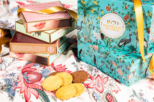 Decorative boxes with floral patterns and a stack of books on a floral surface. Invitation au Voyage Tin – Assorted French Sablés – Maison Holder