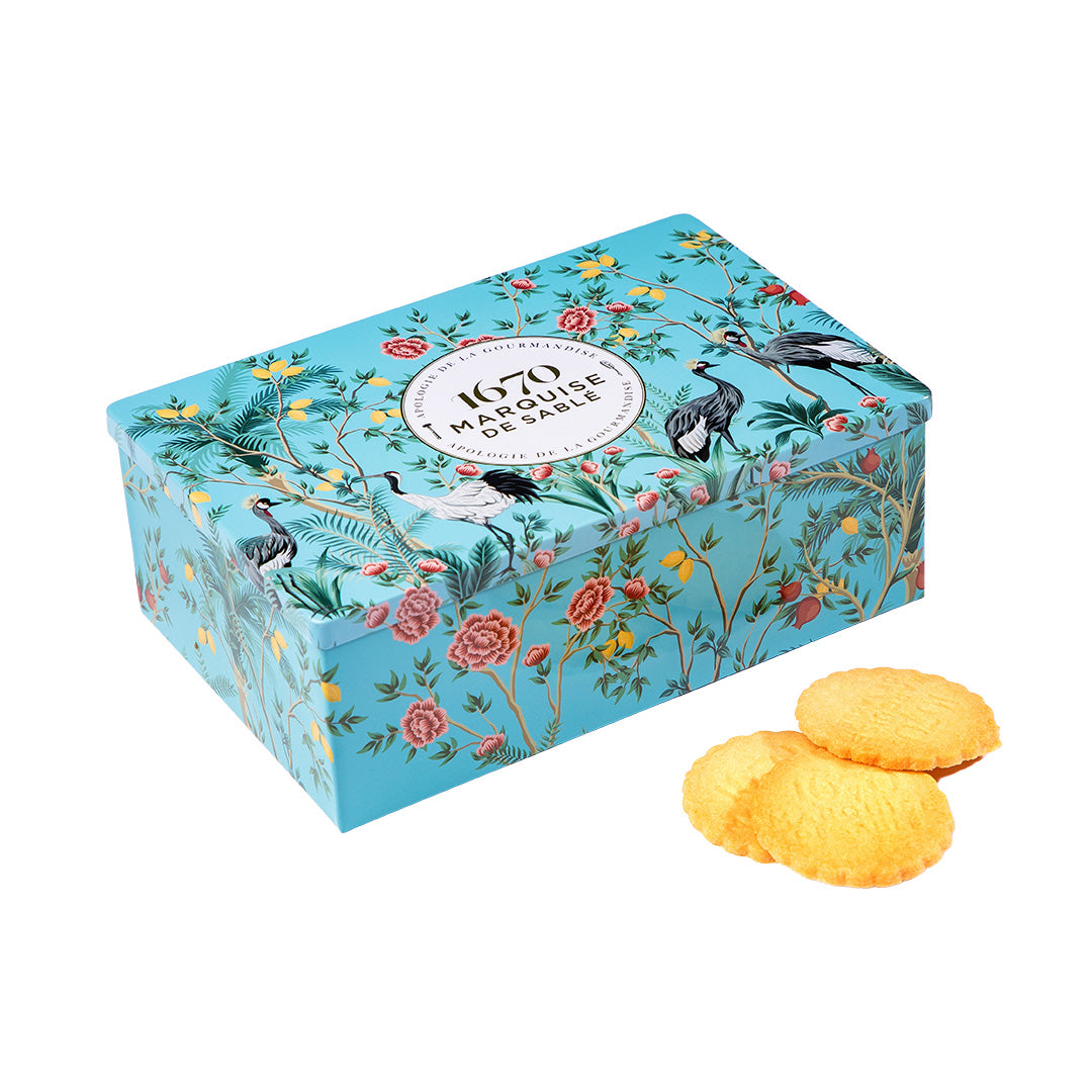 Decorative box with floral and bird design, featuring a brand logo, on a white background with cookies in front. Invitation au Voyage Tin – Assorted French Sablés – Maison Holder