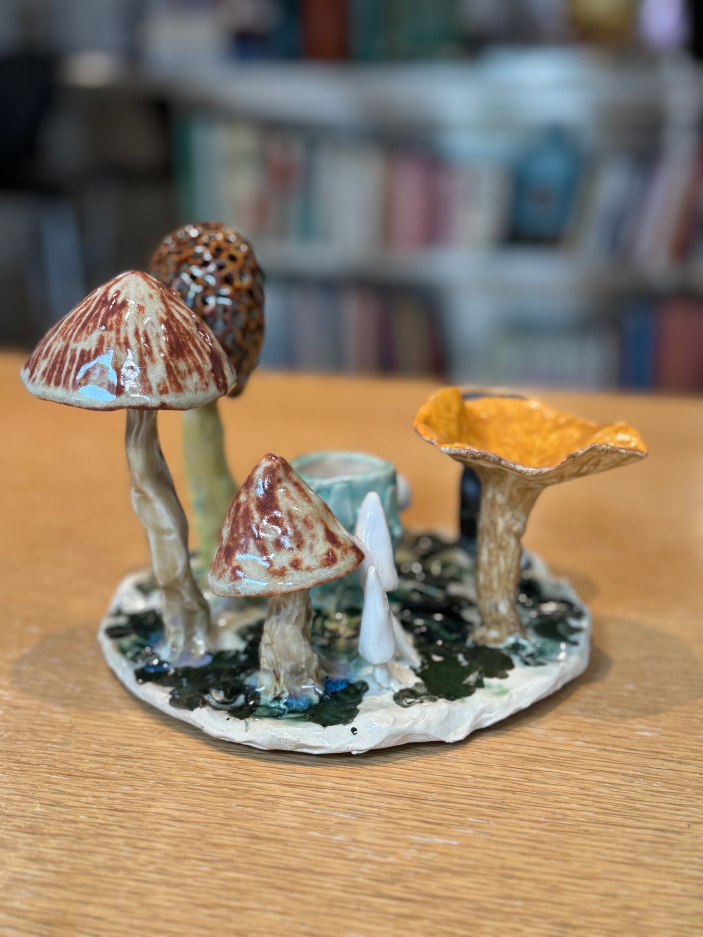 Collection of ceramic mushrooms on a wooden surface with a blurred bookshelf background, Mushroom Candles – Handmade Ceramic – Nathalie Reignault Sann