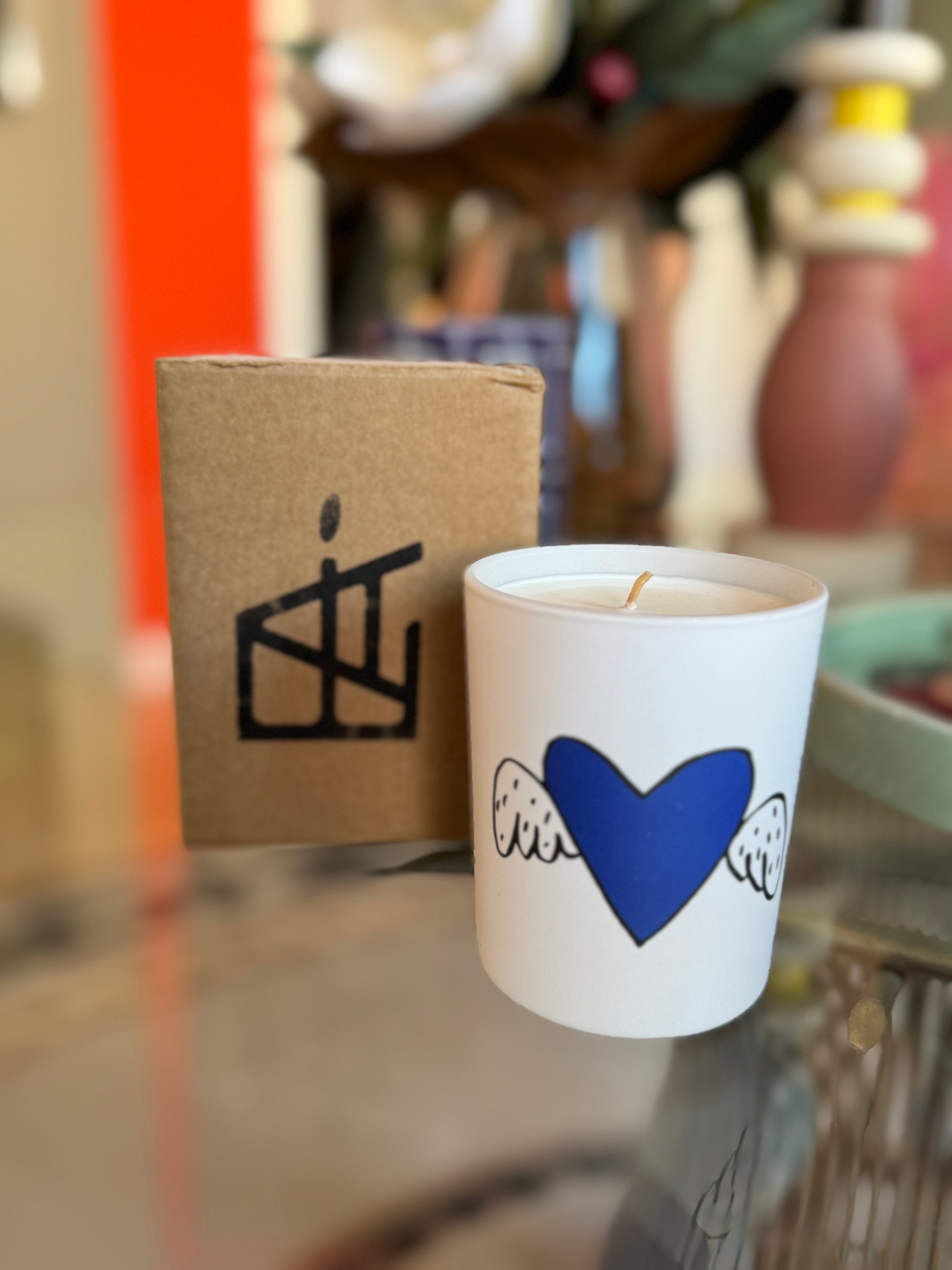 Candle with blue heart design on a white container next to a cardboard box with a black logo. Jean-Charles de Castelbajac Art Candle – Épreuve d’Artistes – Maison Holder