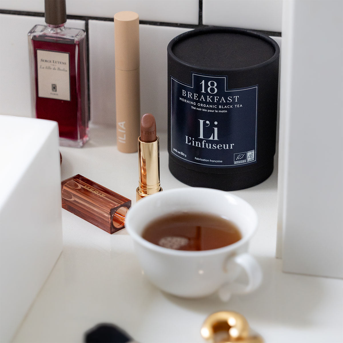 Tea cup with a tea canister, lipstick, and other items on a white surface. Organic Breakfast Tea N°18 – Black Tea Blend by L'infuseur