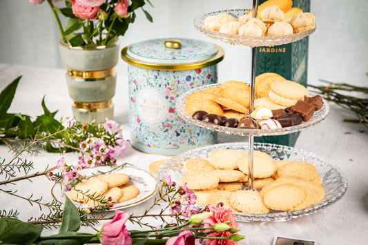 Three-tiered stand with assorted cookies on a table with flowers and decorative tins. Bouquet Sauvage Tin – Butter & Chocolate Sablés – Maison Holder