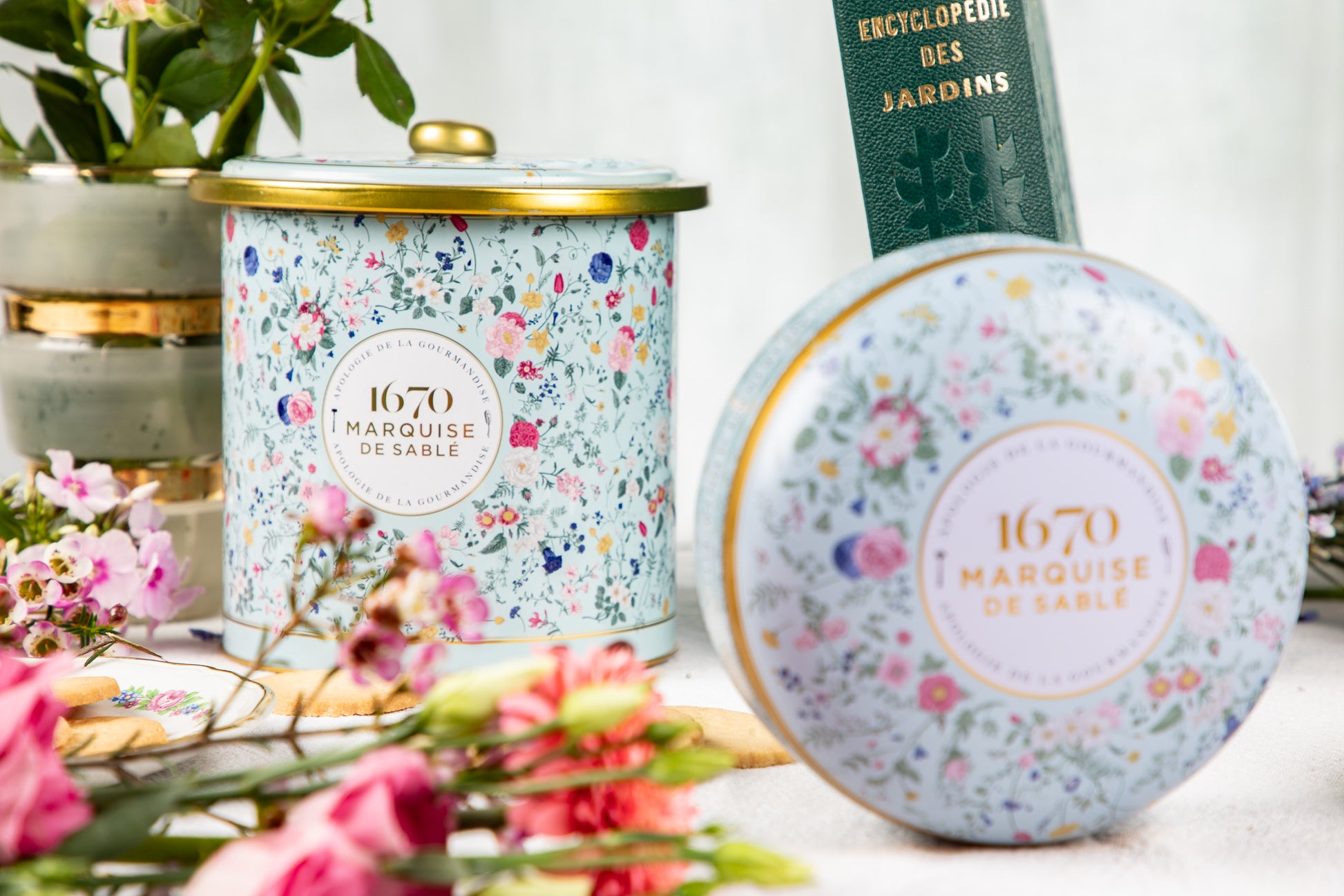 Decorative candle with floral design and '1670 Marquise de Sable' branding, surrounded by flowers and a book. Bouquet Sauvage Tin – Butter & Chocolate Sablés – Maison Holder