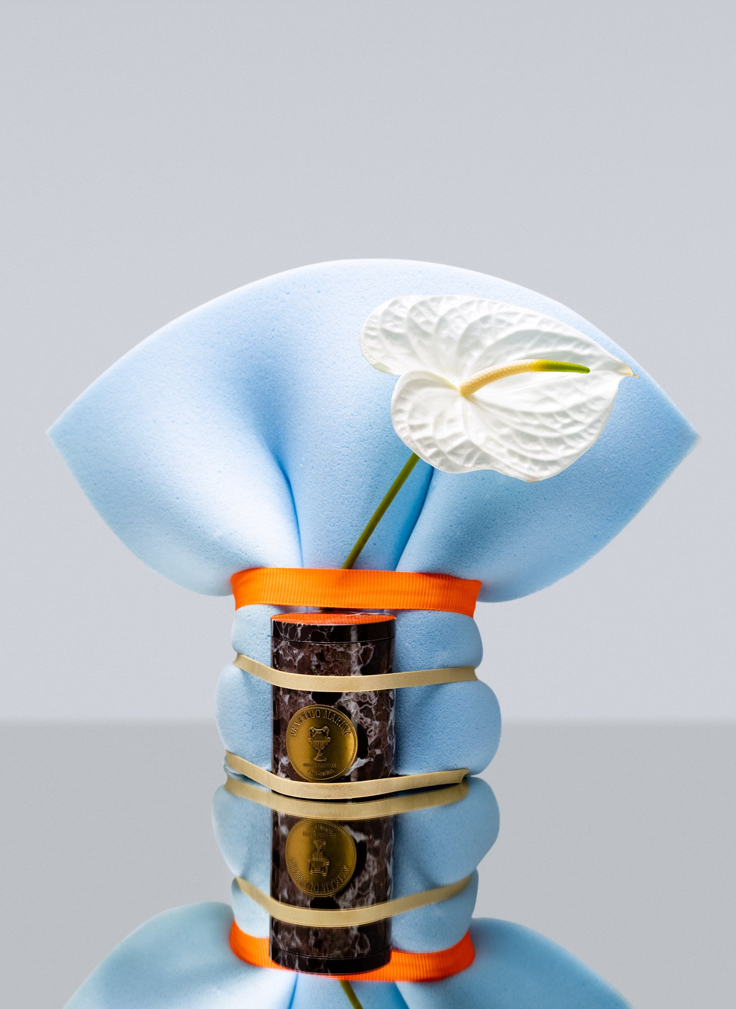 Decorative bottle with a blue wrap, white flower, and orange accents on a light gray background, Lipari Marble Candle – Venetico Marina – Maison Holder