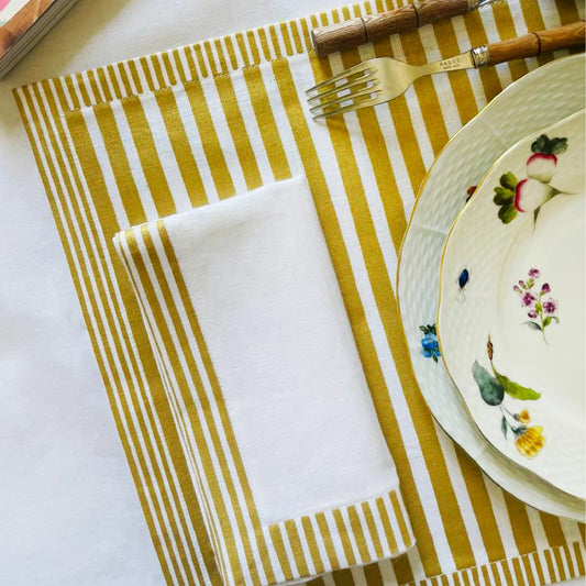 Yellow and white striped placemat with a white napkin, plate, and cutlery on a white surface. Milleraies Striped Linen Napkin – Aurelie Bidermann – Maison Holder