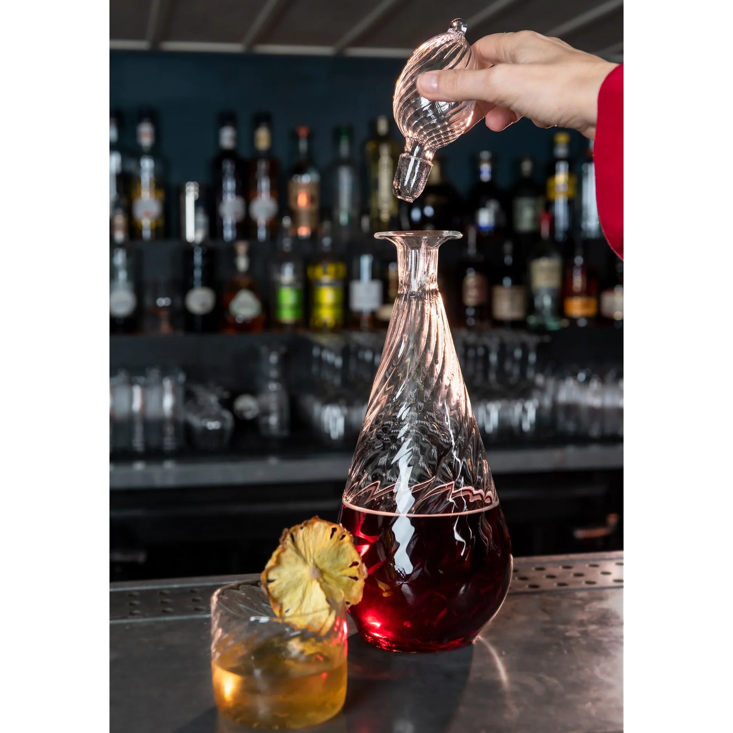 Person pouring a drink from a glass decanter into a glass with a bar setting in the background., Nabucho Wine Carafe – Mouth-Blown Borosilicate Glass – Maison Holder