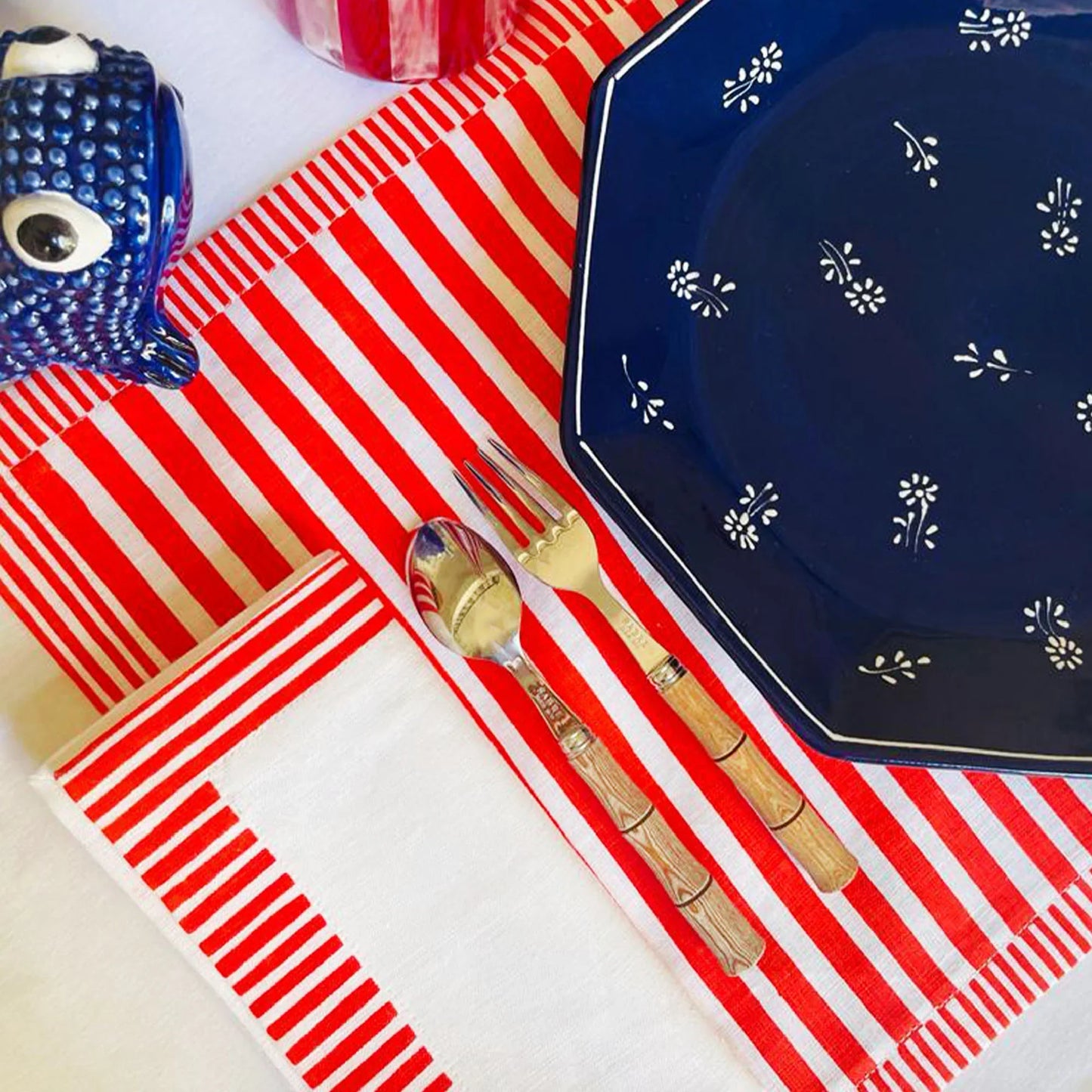 Napkin with red and white stripes, blue plate with white patterns, gold fork, and blue decorative object on a white surface. Milleraies Striped Linen Placemat – Aurelie Bidermann – Maison Holder