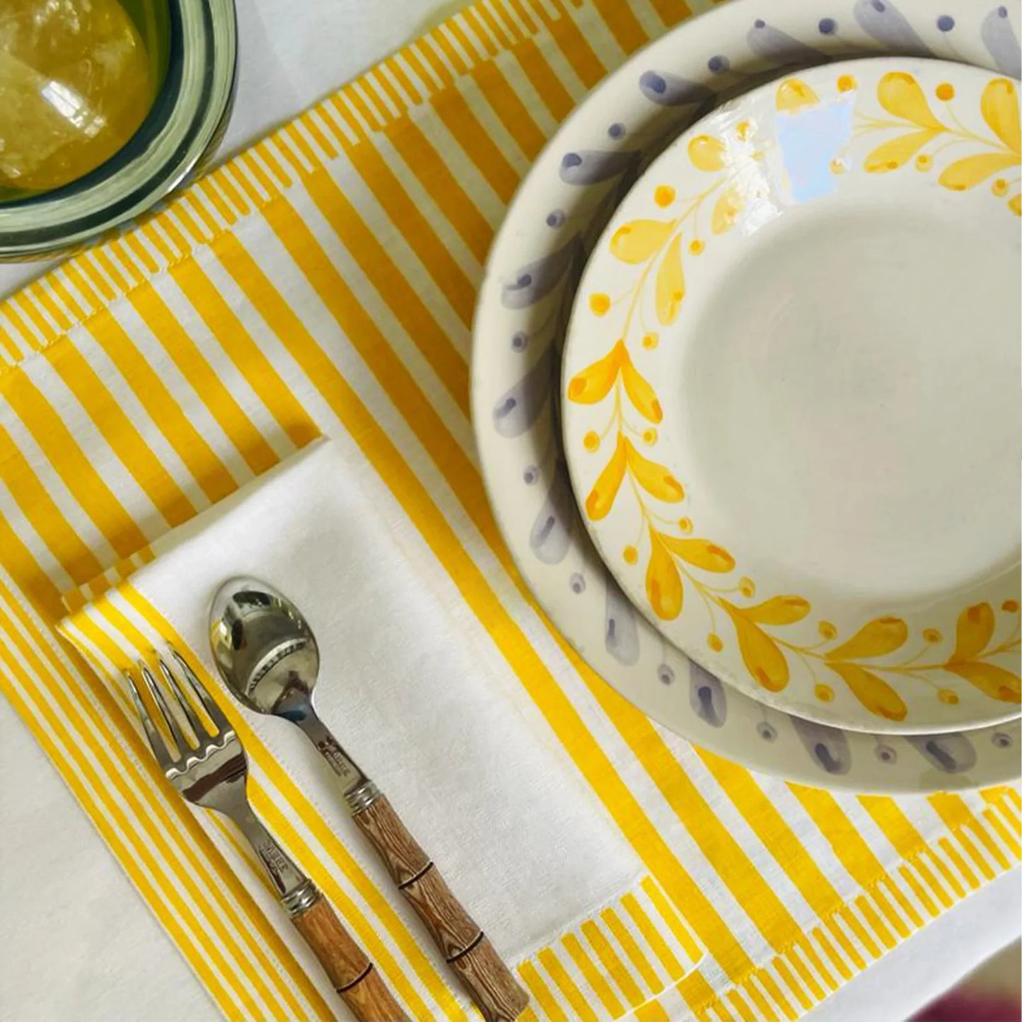 Dinner setting with yellow and white patterned plates, silver fork, and wooden-handled spoon on a yellow and white striped placemat Milleraies Striped Linen Placemat – Aurelie Bidermann – Maison Holder