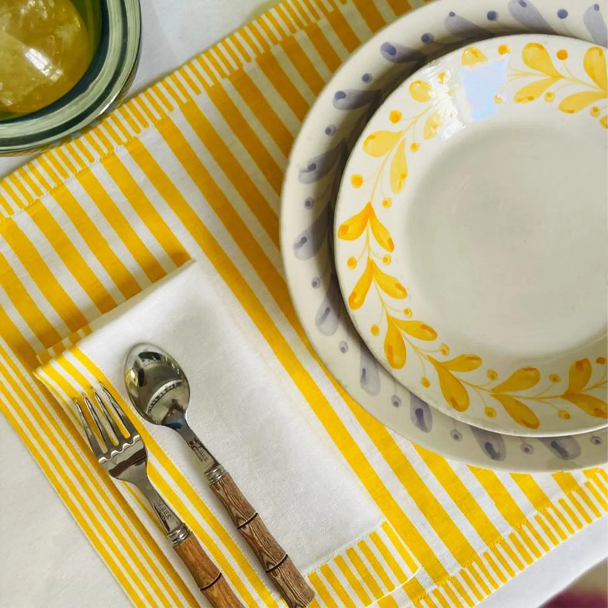 Dinner setting with yellow and white patterned plates, silver fork, and wooden-handled spoon on a yellow and white striped placemat Milleraies Striped Linen Placemat – Aurelie Bidermann – Maison Holder