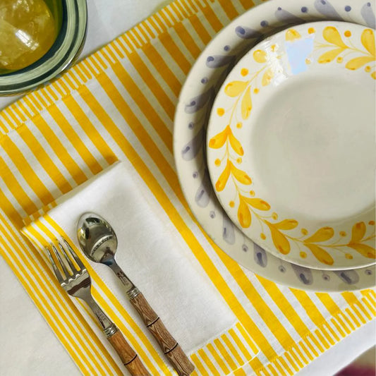 Dinner setting with yellow and white patterned plates, silver fork, and wooden-handled spoon on a yellow and white striped placemat Milleraies Striped Linen Placemat – Aurelie Bidermann – Maison Holder