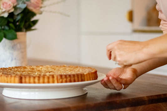 Person reaching for a tart on a wooden table with a blurred background ,Cake Platter Rouen Decor – Desvres Earthenware – Maison Holder