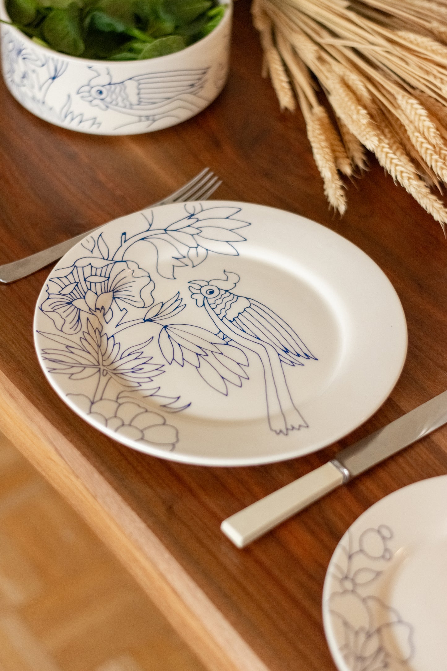 Decorative plate with bird and floral design on a wooden table with cutlery and a bowl. Dinner Plate Rouen Decor – Desvres Earthenware – Maison Holder